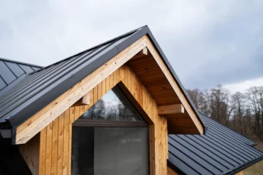 Detail of a modern house roof featuring a wooden gable and dark metal shingles against a cloudy sky.