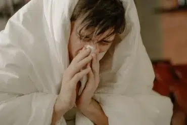 A person wrapped in a white blanket holds a tissue to their nose, appearing unwell. The scene conveys a sense of discomfort and illness.