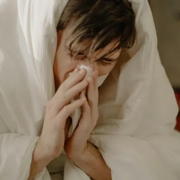 A person wrapped in a white blanket holds a tissue to their nose, appearing unwell. The scene conveys a sense of discomfort and illness.