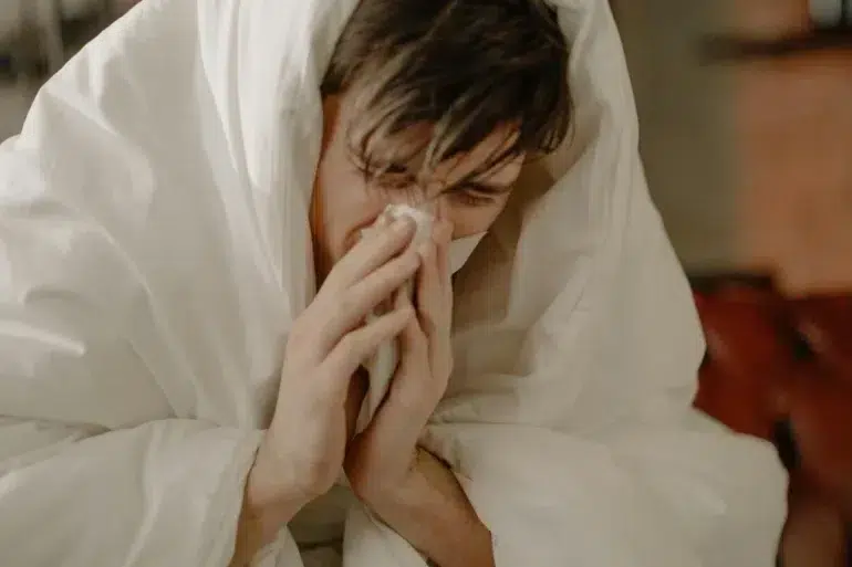 A person wrapped in a white blanket holds a tissue to their nose, appearing unwell. The scene conveys a sense of discomfort and illness.