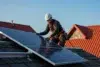 A worker in a safety harness installs solar panels on a tiled roof under a clear blue sky. The scene conveys efficiency and sustainable energy.