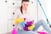 Woman sitting on floor with cleaning supplies. She wears yellow gloves, holding a purple bucket filled with cloths in a bright, tidy room.