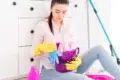 Woman sitting on floor with cleaning supplies. She wears yellow gloves, holding a purple bucket filled with cloths in a bright, tidy room.