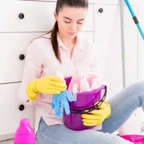 Woman sitting on floor with cleaning supplies. She wears yellow gloves, holding a purple bucket filled with cloths in a bright, tidy room.