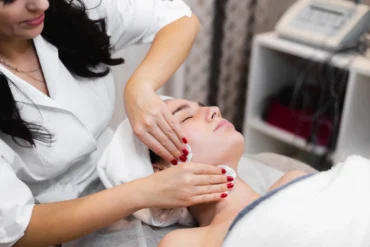 A woman receives a facial massage in a spa setting, lying relaxed with a towel around her head. The therapist wears a white robe, creating a calm atmosphere.