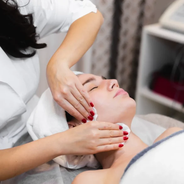 A woman receives a facial massage in a spa setting, lying relaxed with a towel around her head. The therapist wears a white robe, creating a calm atmosphere.