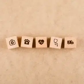 Five wooden blocks featuring health-related icons: a medical bag, house with a cross, heart, stethoscope, and ambulance on a beige background.