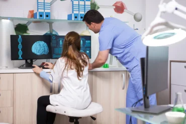 Two healthcare professionals in a lab study brain scans and DNA on dual monitors. The atmosphere is focused, with digital graphics displayed.