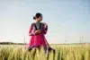 A woman in a vibrant pink and blue traditional dress stands gracefully in a golden wheat field under a clear sky, exuding a serene and elegant aura.