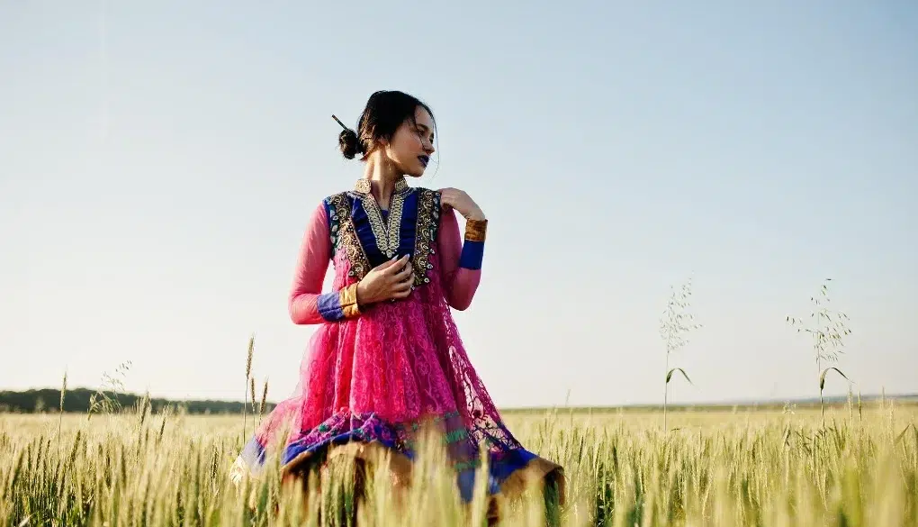 A woman in a vibrant pink and blue traditional dress stands gracefully in a golden wheat field under a clear sky, exuding a serene and elegant aura.