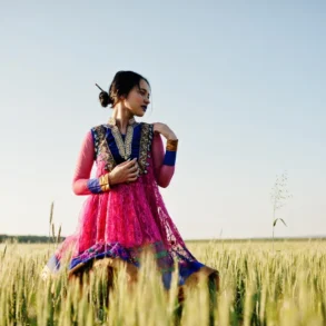 A woman in a vibrant pink and blue traditional dress stands gracefully in a golden wheat field under a clear sky, exuding a serene and elegant aura.