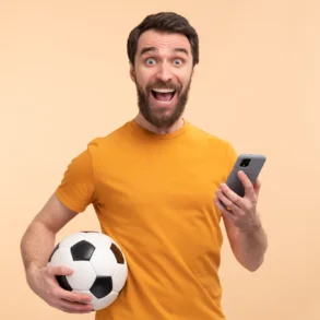 A bearded man in a yellow shirt, excitedly holding a soccer ball and smartphone against a peach background, conveys happiness and enthusiasm.
