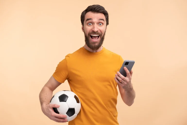 A bearded man in a yellow shirt, excitedly holding a soccer ball and smartphone against a peach background, conveys happiness and enthusiasm.