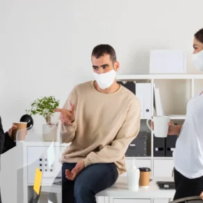Three people wearing masks engage in conversation in an office. One sits on a desk, gesturing. Two others stand with coffee, promoting a relaxed atmosphere.