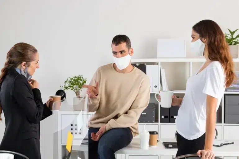 Three people wearing masks engage in conversation in an office. One sits on a desk, gesturing. Two others stand with coffee, promoting a relaxed atmosphere.