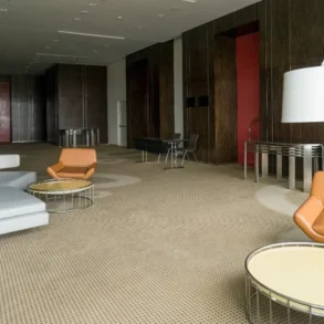 Modern lounge with a beige carpet, brown wood-paneled walls, orange and gray chairs, circular tables, and a large white floor lamp. Calm atmosphere.