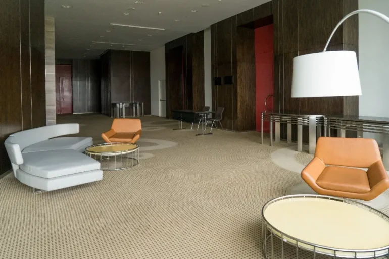 Modern lounge with a beige carpet, brown wood-paneled walls, orange and gray chairs, circular tables, and a large white floor lamp. Calm atmosphere.