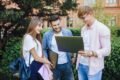 Young adults stand in a park, smiling and looking at a laptop. They are casually dressed and appear happy, engaged in a group conversation.