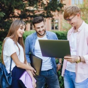 Young adults stand in a park, smiling and looking at a laptop. They are casually dressed and appear happy, engaged in a group conversation.
