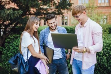 Young adults stand in a park, smiling and looking at a laptop. They are casually dressed and appear happy, engaged in a group conversation.