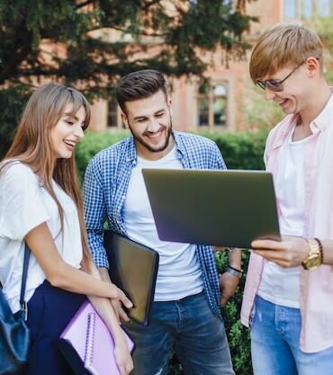 Young adults stand in a park, smiling and looking at a laptop. They are casually dressed and appear happy, engaged in a group conversation.