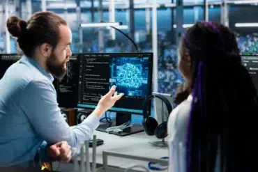 A man and woman discuss artificial intelligence at a workstation. The monitor displays a digital brain. The setting feels collaborative and modern.