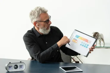 Elderly man with glasses and earbuds points to a colorful chart on a clipboard. A retro clock shows Saturday 22. Calm, focused office setting.