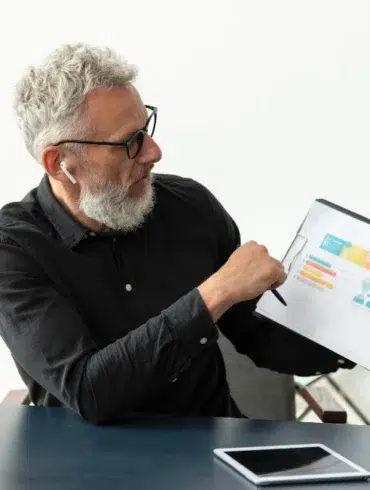 Elderly man with glasses and earbuds points to a colorful chart on a clipboard. A retro clock shows Saturday 22. Calm, focused office setting.