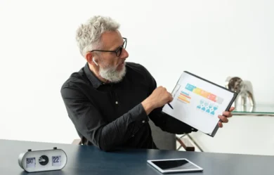 Elderly man with glasses and earbuds points to a colorful chart on a clipboard. A retro clock shows Saturday 22. Calm, focused office setting.
