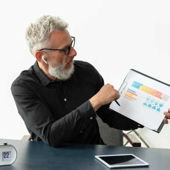 Elderly man with glasses and earbuds points to a colorful chart on a clipboard. A retro clock shows Saturday 22. Calm, focused office setting.