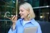 A smiling woman with blonde hair holds a smartphone, speaking into it. She wears a blue shirt and clutches a closed laptop. Office building in the background.
