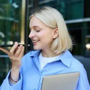 A smiling woman with blonde hair holds a smartphone, speaking into it. She wears a blue shirt and clutches a closed laptop. Office building in the background.