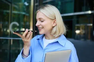 A smiling woman with blonde hair holds a smartphone, speaking into it. She wears a blue shirt and clutches a closed laptop. Office building in the background.