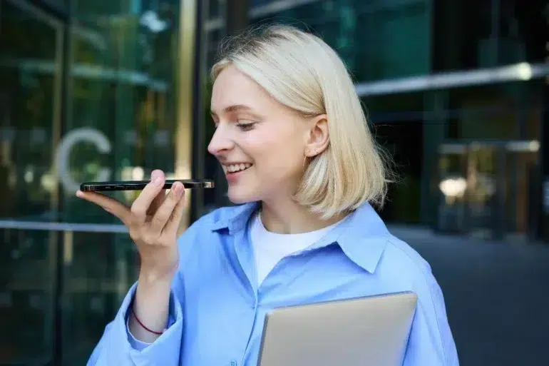 A smiling woman with blonde hair holds a smartphone, speaking into it. She wears a blue shirt and clutches a closed laptop. Office building in the background.