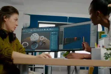 Two women collaborate at a desk with dual monitors displaying 3D design software. The setting is a modern office, with a focused, creative atmosphere.