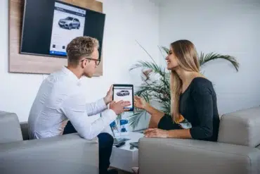 A man and woman sit in a modern office discussing a car on a tablet, smiling. A digital car display is on the wall. The atmosphere is professional yet friendly.