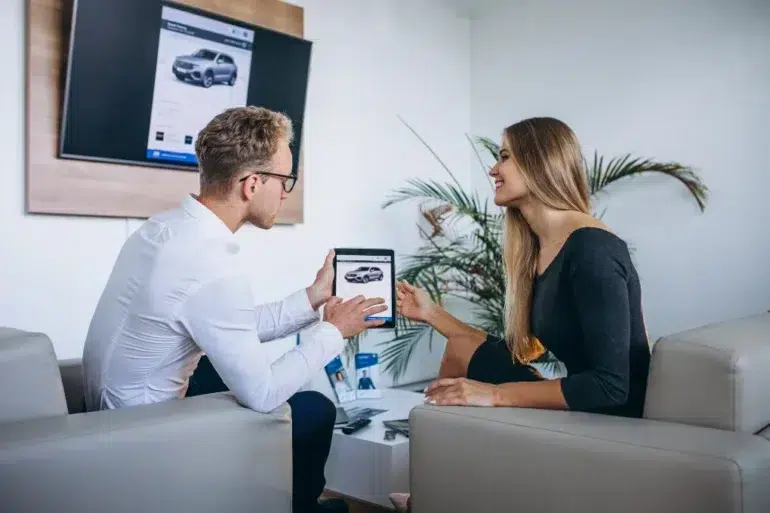 A man and woman sit in a modern office discussing a car on a tablet, smiling. A digital car display is on the wall. The atmosphere is professional yet friendly.