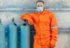 Man in an orange jumpsuit and protective gear stands confidently next to blue gas cylinders against a weathered wall, conveying safety and professionalism.