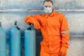 Man in an orange jumpsuit and protective gear stands confidently next to blue gas cylinders against a weathered wall, conveying safety and professionalism.