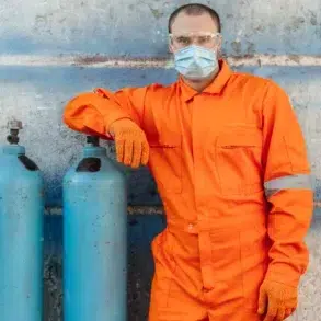 Man in an orange jumpsuit and protective gear stands confidently next to blue gas cylinders against a weathered wall, conveying safety and professionalism.