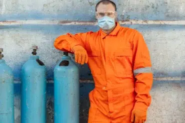 Man in an orange jumpsuit and protective gear stands confidently next to blue gas cylinders against a weathered wall, conveying safety and professionalism.
