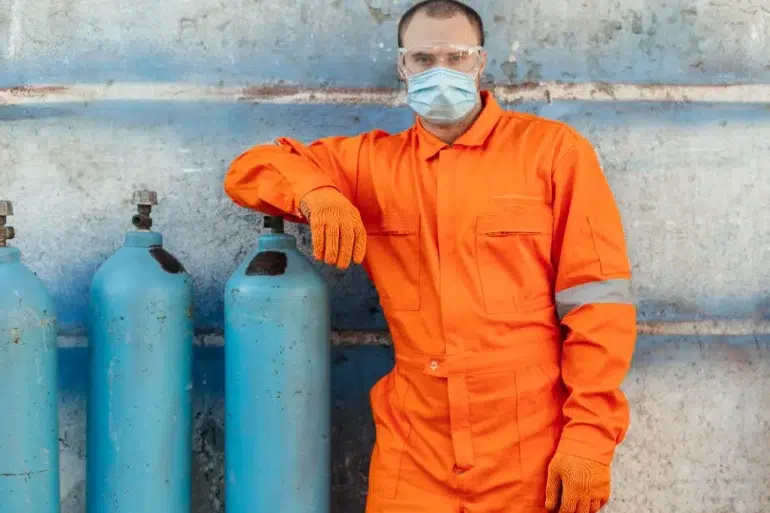 Man in an orange jumpsuit and protective gear stands confidently next to blue gas cylinders against a weathered wall, conveying safety and professionalism.