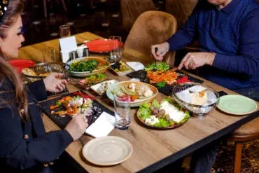 Two people dining at a wooden table, enjoying a variety of colorful dishes, including salads, olives, and appetizers, creating a lively, cozy atmosphere.