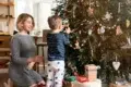 A woman and a child decorate a Christmas tree. The child reaches to hang an ornament while the woman kneels beside him, smiling warmly. Presents are beneath the tree.