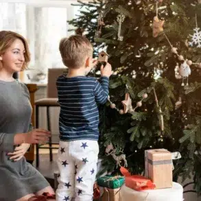 A woman and a child decorate a Christmas tree. The child reaches to hang an ornament while the woman kneels beside him, smiling warmly. Presents are beneath the tree.