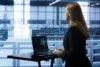 A woman in a black suit stands at a desk with a laptop, looking at data center servers. The atmosphere is professional and focused.