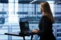 A woman in a black suit stands at a desk with a laptop, looking at data center servers. The atmosphere is professional and focused.