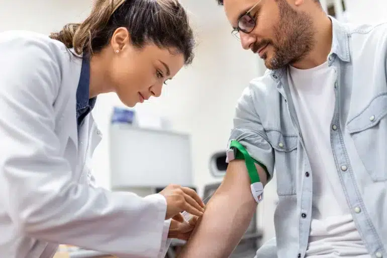 A healthcare professional draws blood from a man in a clinical setting. The man is seated and has a tourniquet on his arm. Both appear focused and calm.