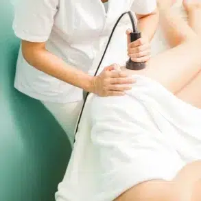 A person in a white uniform uses a device on a client's leg, covered with a towel, suggesting a medical or spa treatment, conveying relaxation and care.