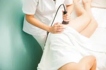 A person in a white uniform uses a device on a client's leg, covered with a towel, suggesting a medical or spa treatment, conveying relaxation and care.